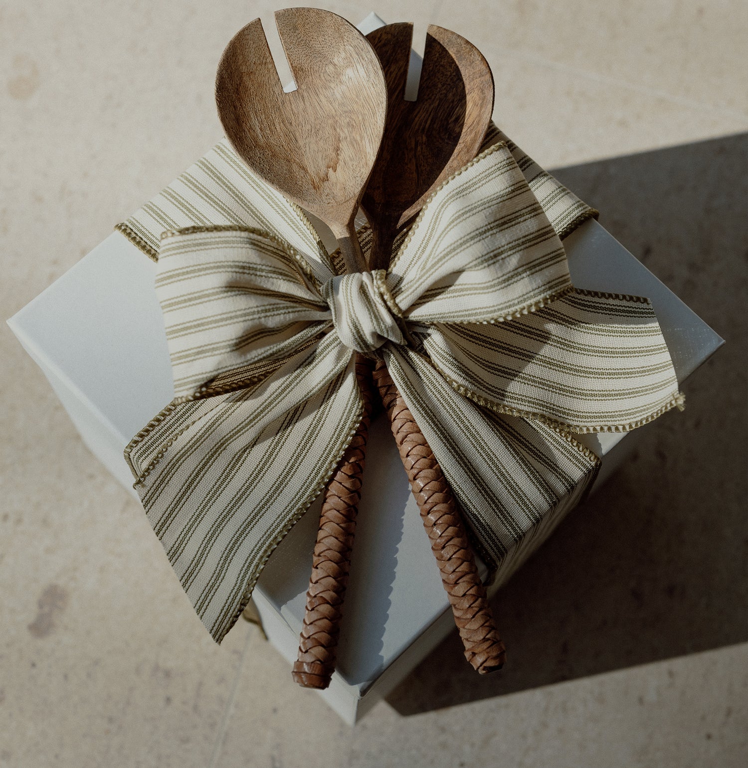 White box with cream and green striped ribbon wrapped in a bow, with wooden spoons held within the knot on top of the box. 